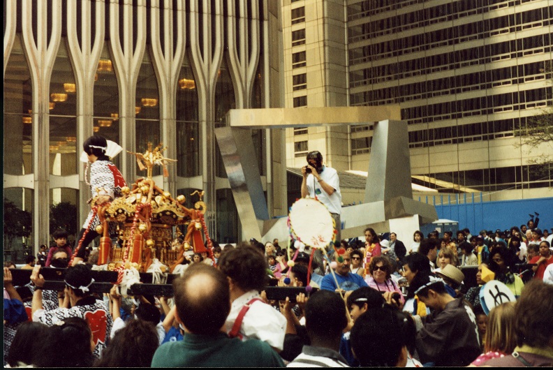 A large crowd of people in front of a Twin Tower. A man stands in front of an abstract steel sculpture of four intersecting beams taking a photograph of a procession carrying a pagoda-like object in red and gold.