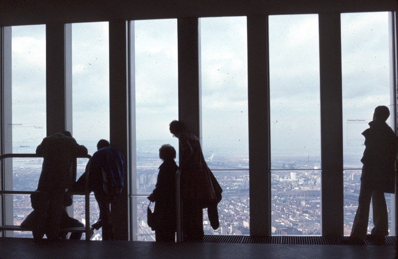 Silhouette shot of five visitors in front of a row of six full length windows, looking down to a bird's eye view of the city on a cloudy day.
