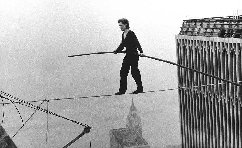 Tightrope walker holding a pole and walking across a rope with the top of one Twin Tower visible on the right in the black and white photograph.